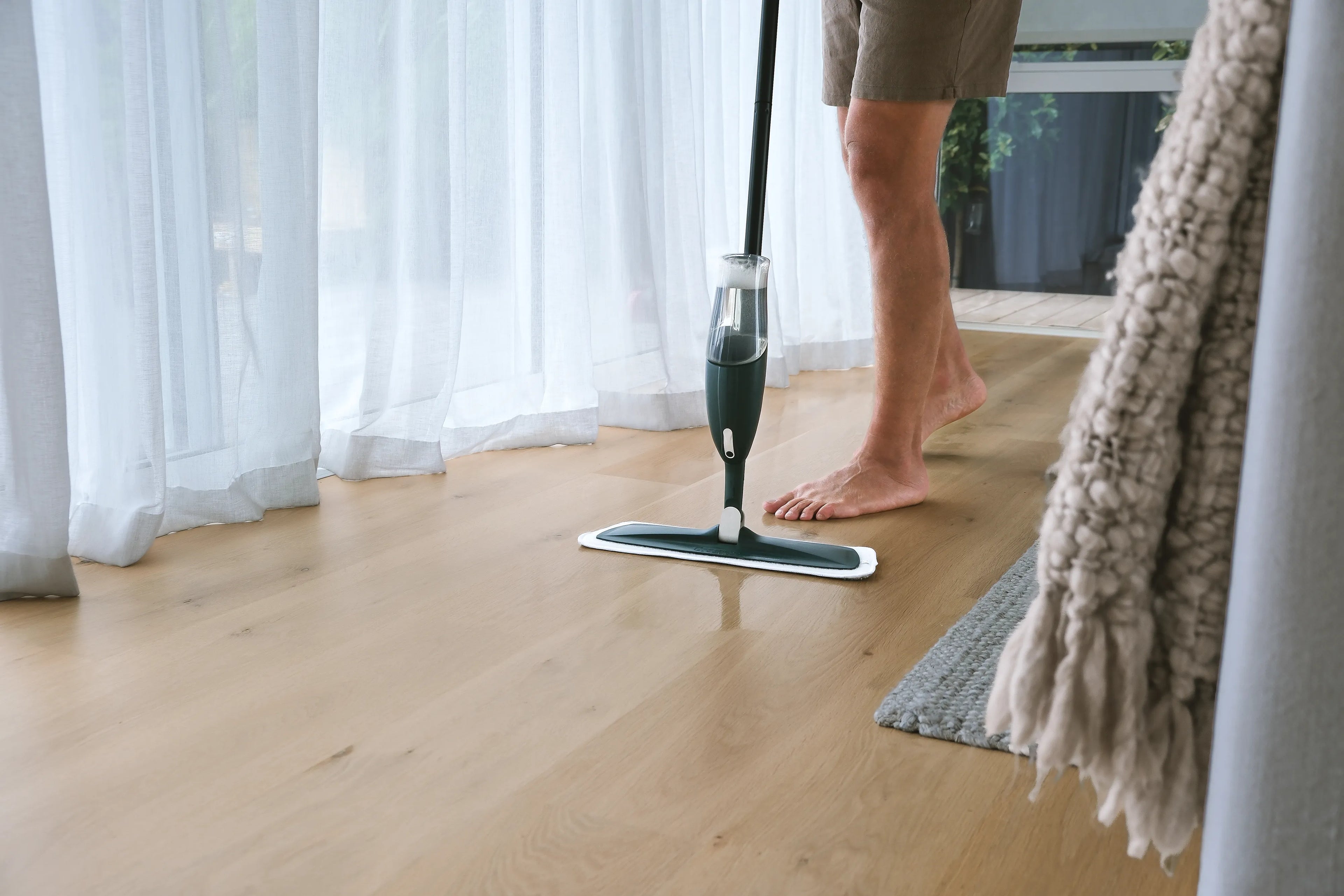 Person using a floor mop on a wooden floor with white curtains in the background