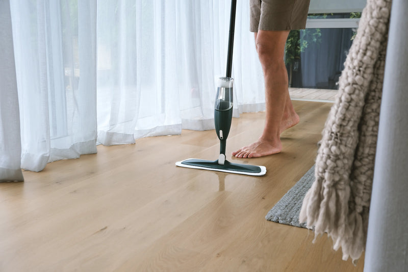 Person using a floor mop on a wooden floor with white curtains in the background